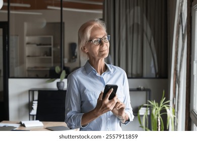 Pensive middle-aged businesswoman wear eyeglasses holding smartphone using mobile business application, researching market trends, coordinating tasks, making calls. Workflow using modern technology - Powered by Shutterstock