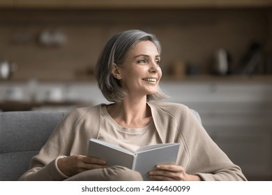 Pensive middle-aged woman relaxing in living room with book, sitting on cozy sofa, read favorite bestseller or novel, looking away, thinking over story, dreaming, enjoy literature hobby alone at home