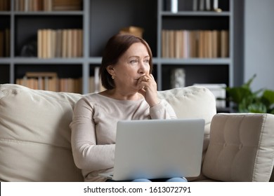 Pensive middle-aged woman sit on couch in living room using laptop look in distance thinking or pondering, thoughtful senior female distracted lost in thoughts feel lonely or sad at home