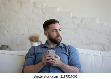 Pensive Millennial Bearded Single Man Consider Purchase Sit On Sofa Hold Smartphone Distracted From Gadget Usage Staring Aside. Modern Tech, Remote Communication, Leisure Using Technologies Concept