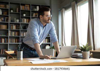 Pensive millennial Caucasian male employee stand at home office desk look in distance think ponder over problem solution. Thoughtful man distracted from computer work make plan decision at workplace.