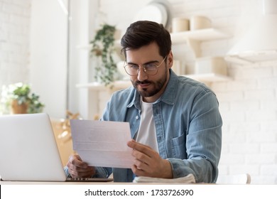 Pensive Millennial Man In Glasses Sit At Table Work On Laptop Read Letter Correspondence Thinking, Thoughtful Young Male Distracted From Computer Consider Paperwork Or Post Notification At Home