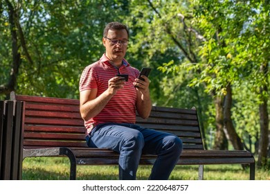 Pensive Millennial Man In Glasses Using Smartphone And Credit Card, Sitting On Wooden Bench In Sunny City Park. Confident Caucasian Man Purchasing Online On Cellphone