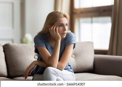 Pensive millennial woman sit on couch in living room look in distance thinking pondering, thoughtful Caucasian young female relax on sofa at home lost in thoughts, planning or visualizing