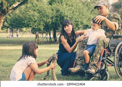 Pensive Mom And Disabled Military Dad In Wheelchair Spending Leisure Time With Kids Outdoors, Arranging Fuelwood For Fire On Grass. Disabled Veteran Or Family Outdoors Concept