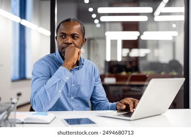 Pensive office worker in blue shirt thinking at desk with open laptop and documents. Modern office environment with bright lights and large windows. Concept of contemplation, problem-solving - Powered by Shutterstock