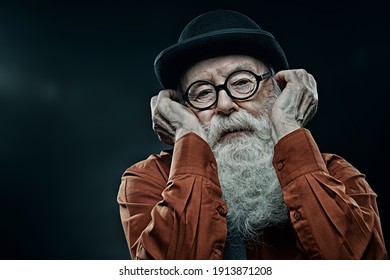 Pensive old man with long white beard in bowler hat and glasses looking at the camera. Old age concept. Black background with copy space.