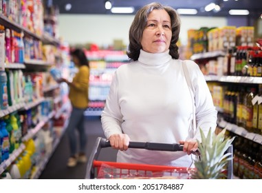 Pensive Older Woman Shopping At Store, Walking With Cart Among Shelves And Choosing Products