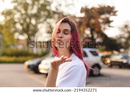 Pensive pink hair woman turn side and make blow kisses walking on the street. Outdoor shot of happy hippie lady in boho freedom style. Girl send air kiss to camera, closed eyes, love concept.