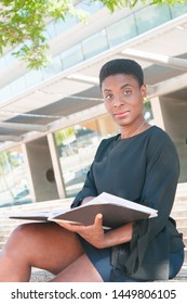 Pensive Positive Black Woman Studying Album Outside. African American Lady Sitting On Outdoor Stair, Holding Photo Book And Looking At Camera. Book Reader Concept