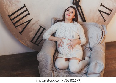 Pensive pregnant woman dreaming about child sitting in armchair with huge clock behind - Shutterstock ID 763301179