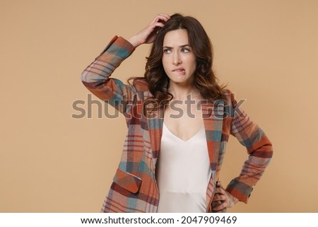 Pensive preoccupied confused young brunette woman 20s in casual checkered jacket posing standing put hand on head biting lips looking aside isolated on pastel beige colour background, studio portrait