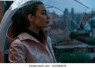 Pensive Pretty Girl Holding Transparent Umbrella While Strolling Outside On Rainy Autumn Day. Photo Looks Like Film Frame Or Movie Screenshot. Processed In Cold Colors