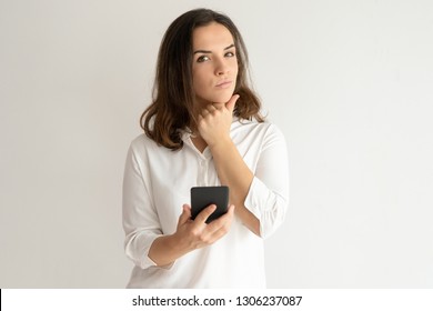 Pensive pretty young woman holding smartphone. Lady thinking, touching chin and looking at camera. Technology and contemplation concept. Isolated view on white background. - Shutterstock ID 1306237087