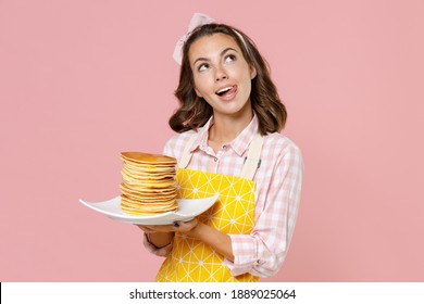 Pensive Pretty Young Woman Housewife In Yellow Apron Hold Plate With Pancakes Looking Up Licking Lips Doing Housework Isolated On Pastel Pink Colour Background Studio Portrait. Housekeeping Concept