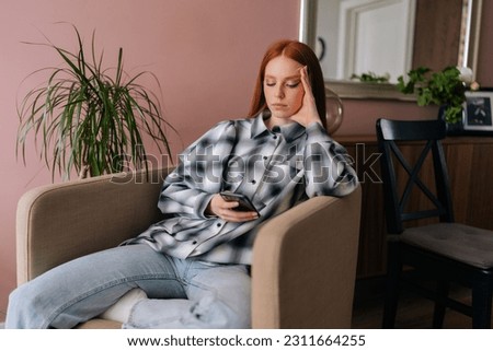 Pensive pretty young woman relaxing at home sitting in armchair, thoughtful looking to smartphone screen, using online app. Serious young woman in casual clothes messaging on mobile phone at home.