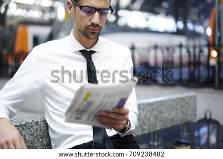 Pensive proud ceo in spectacles reading newspaper articles while waiting for public transport on subway, serious male in formal wear checking latest news from press sitting near copy space