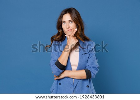 Pensive puzzled perplexed confused concerned young brunette woman 20s in basic jacket standing put hand prop up on chin looking camera isolated on bright blue colour wall background, studio portrait
