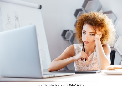 Pensive redhead curly young woman designer using laptop and grapic tablet on workplace  - Powered by Shutterstock