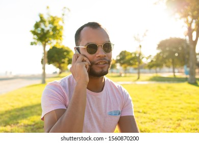 Pensive Relaxed Handsome Guy Talking On Mobile Phone Outdoors. Latin Man Sitting On Grass In Park, Speaking On Cellphone. Phone Call Concept