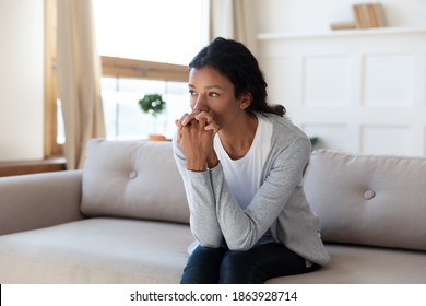 Pensive sad young African American woman sit on sofa at home look in distance pondering thinking. Unhappy upset millennial female suffer from depression or solitude, feel lonely. Miscarriage concept.