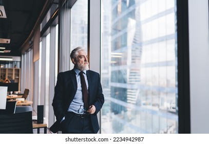 Pensive senior male CEO with gray hair in classy clothes holding eyeglasses and looking through window while working in modern office - Powered by Shutterstock