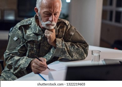 Pensive Senior Veteran In Army Uniform Sitting At Table In The Office And Filling Documents. 