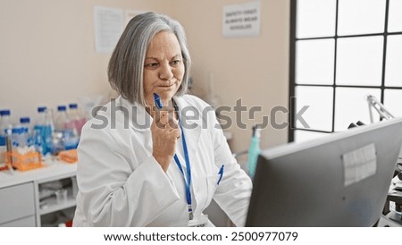 Pensive senior woman scientist in lab with computer, pensively holding pen, indoor