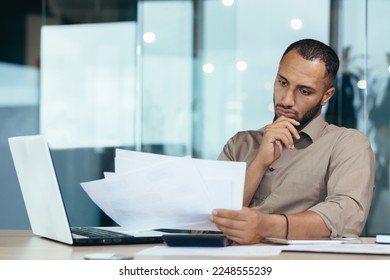 Pensive serious businessman reading financial report, hispanic businessman holding document in hands looking disappointed, working inside modern office with laptop behind paper work. - Powered by Shutterstock