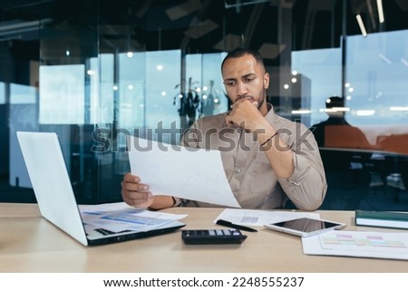 Pensive serious businessman reading financial report, hispanic businessman holding document in hands looking disappointed, working inside modern office with laptop behind paper work.