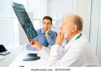 Pensive Serious Mature Adult Male Physician Consult Shocked Young Man Patient Giving Bad News Explaining Results Of MRI Image. Sad Scared Young Man Listening To Bad News Sitting In Doctor Office.