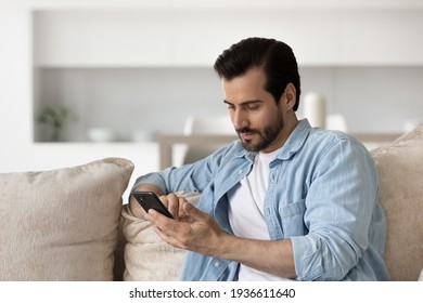 Pensive serious young man sit on sofa at home text message on modern smartphone gadget. Focused millennial male look at cellphone screen browse surf wireless internet on device. Technology concept.