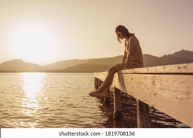 Pensive single Caucasian pretty young woman sitting on a pier of a lake looking down to the water at sunset - Vintage orange color mood - Shutterstock ID 1819708136