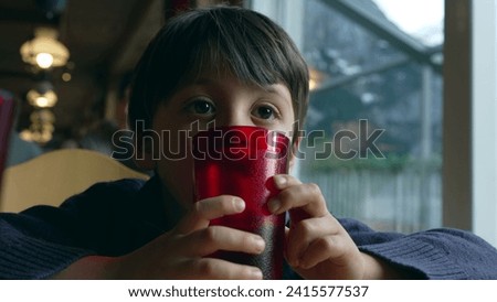 Pensive small boy daydreaming at restaurant leaning on red plastic cup while staring blankly, thoughtful child awaiting food at diner by window