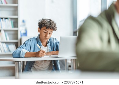 Pensive, smart schoolboy taking notes learning languages, preparing for exam using laptop in modern classroom. Serious brunette guy wearing blue shirt doing homework. Back to school, education concept - Powered by Shutterstock