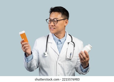 Pensive Smiling Middle Aged Chinese Male Doctor In White Coat, Glasses Looks At Jars Of Pills, Isolated On Blue Background, Studio. Choice Of Medicines, Ad And Offer, Treatment Of Ill And Health Care