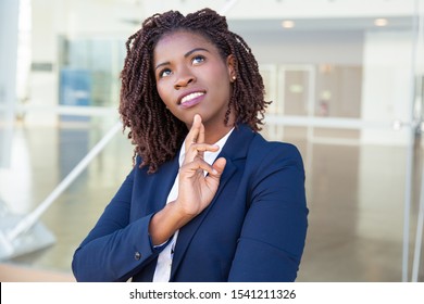 Pensive Smiling Professional Thinking Outside. Young Black Business Woman Standing At Outdoor Glass Wall, Touching Chin And Looking Up. Thinking Concept