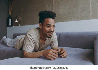 Pensive smiling wistful brunette young african american man 20s wearing casual beige t-shirt sweatpants lay down on grey sofa indoors apartment resting on weekends staying at home during quarantine
