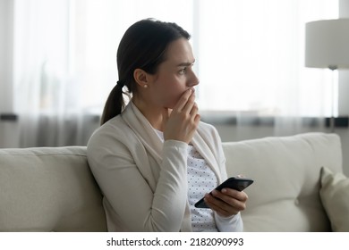 Pensive stressed young Caucasian woman sit on sofa at home using cellphone look in distance pondering of problem. Anxious millennial girl feel distressed frustrated with message or text on smartphone.