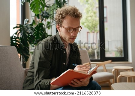Pensive student studying, learning language sitting at workplace.  Education concept. Portrait of young handsome man writer wearing stylish eyeglasses taking notes working from home  