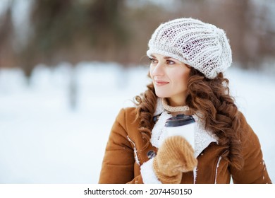 Pensive Stylish Middle Aged Woman With Mittens And Cup Of Hot Beverage In A Knitted Hat And Sheepskin Coat Outside In The City Park In Winter.