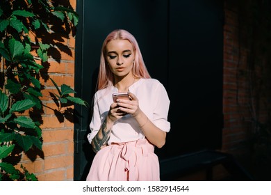 Pensive Stylish Woman With Long Pink Hairs And Tattoo On Hand Surfing Mobile Phone Nearby Black Door Of Bricked Building 