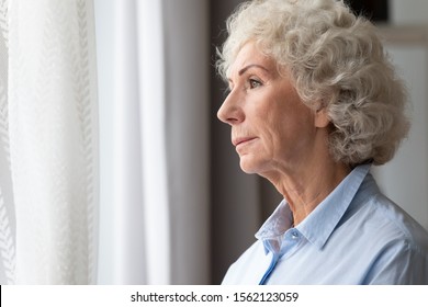 Pensive Thoughtful Lonely Senior Adult Grandma Stand Alone At Home Looking Through Window Waiting, Serious Sad Old Elderly Woman Think Of Loneliness Future Feel Worried Depressed Melancholic Concept