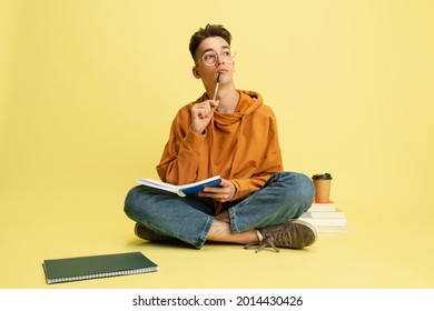 Pensive, thoughtful. One young caucasian guy, student in glasses sits on floor with notepad and books on yellow studio background. Education, studying and student life concept. - Powered by Shutterstock