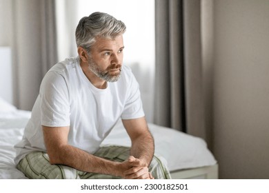 Pensive thoughtful unhappy handsome grey-haired bearded mature caucasian man wearing pajamas sitting on bed at home, looking at copy space, feeling lonely, going through divorce - Powered by Shutterstock