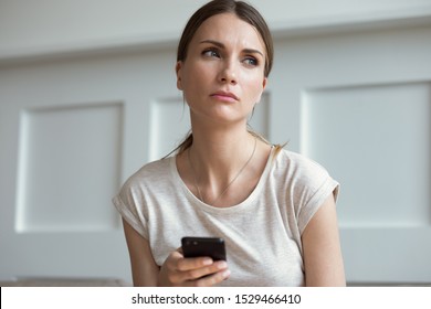 Pensive thoughtful young 30s woman holding smartphone look away lost on sad thoughts waiting first step from man, call or text message or date invitation from boyfriend feels jealousy and concerned