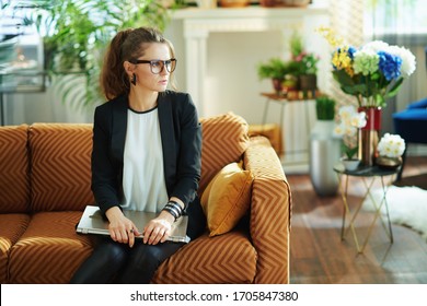 Pensive Trendy Housewife In White Blouse And Black Jacket With Closed Laptop Sitting On Couch At Modern Home In Sunny Day.