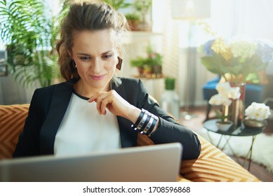 Pensive Trendy Middle Age Woman In White Blouse And Black Jacket At Modern Home In Sunny Day Surfing Web On A Laptop While Sitting On Couch.