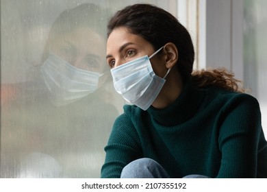 Pensive Unhappy Young Woman In Facial Protective Disposal Mask Sitting On Windowsill, Feeling Lonely During Quarantine Pastime, Sick With Covid19 Disease, Coronavirus Pandemic Illness Concept.