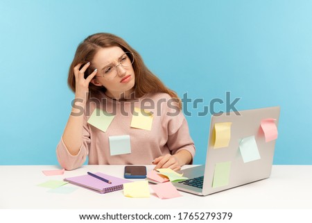 Pensive upset woman employee in nerd eyeglasses sitting at workplace office, all covered with sticky notes and thinking intensely, frustrated by workload. indoor studio shot isolated blue background